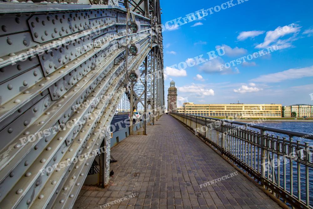 Фотография захватывает перспективу одного из железных мостов Санкт-Петербурга, со стальными балками и креплениями на переднем плане, что подчеркивает инженерную сложность и красоту его конструкции. Вдалеке виднеется красивый исторический башенный часовой здание, а река Нева протекает под мостом, добавляя динамику композиции. Такое изображение подходит для публикаций, посвящённых инженерному искусству, градостроительству и архитектуре города, а также может быть использовано в качестве образовательного материала или декоративного элемента.